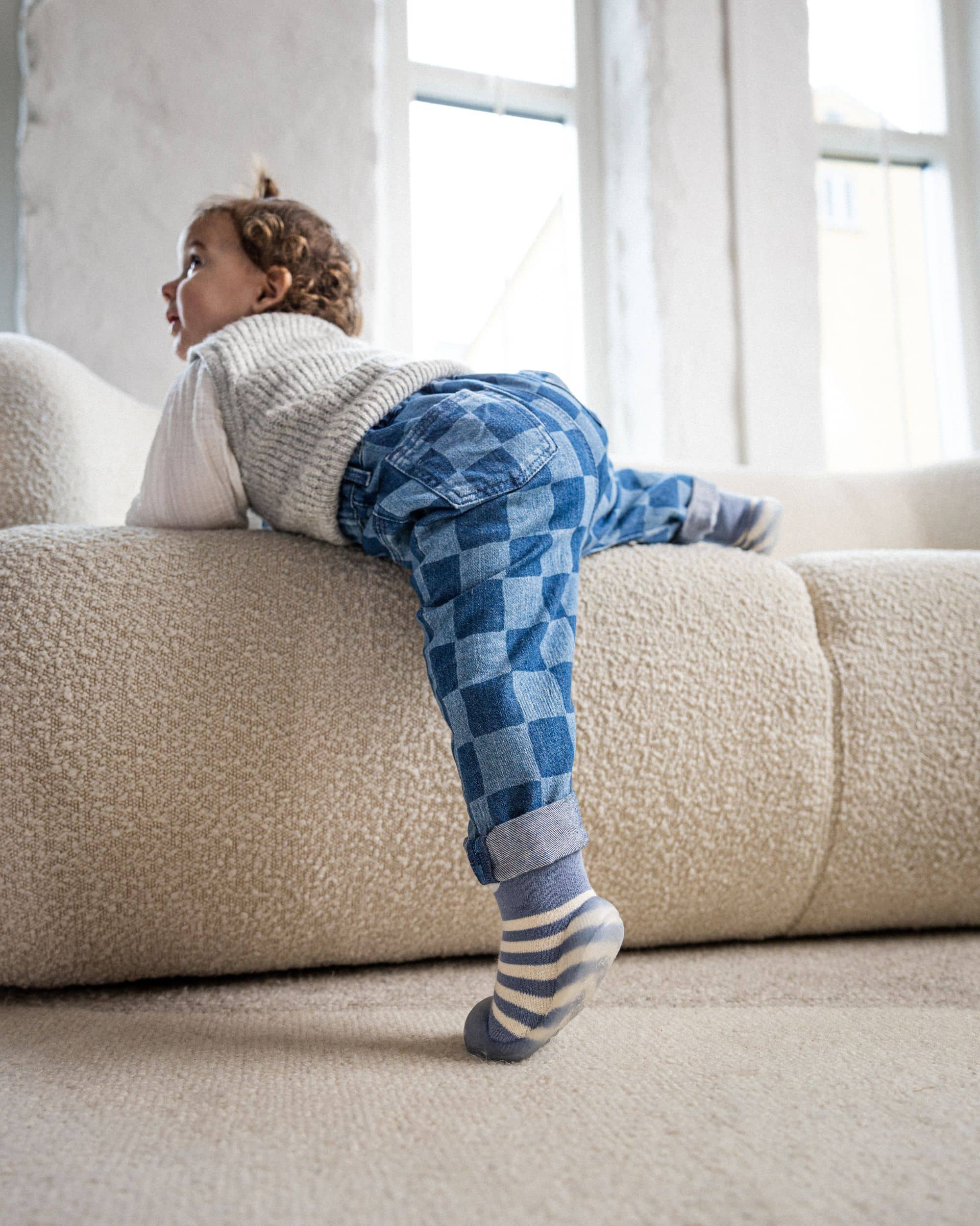 Barn i blå-stribede begyndersko fra Minifus klatrer op ad sofa, iført ternede bukser og strikvest i en lys stue.
