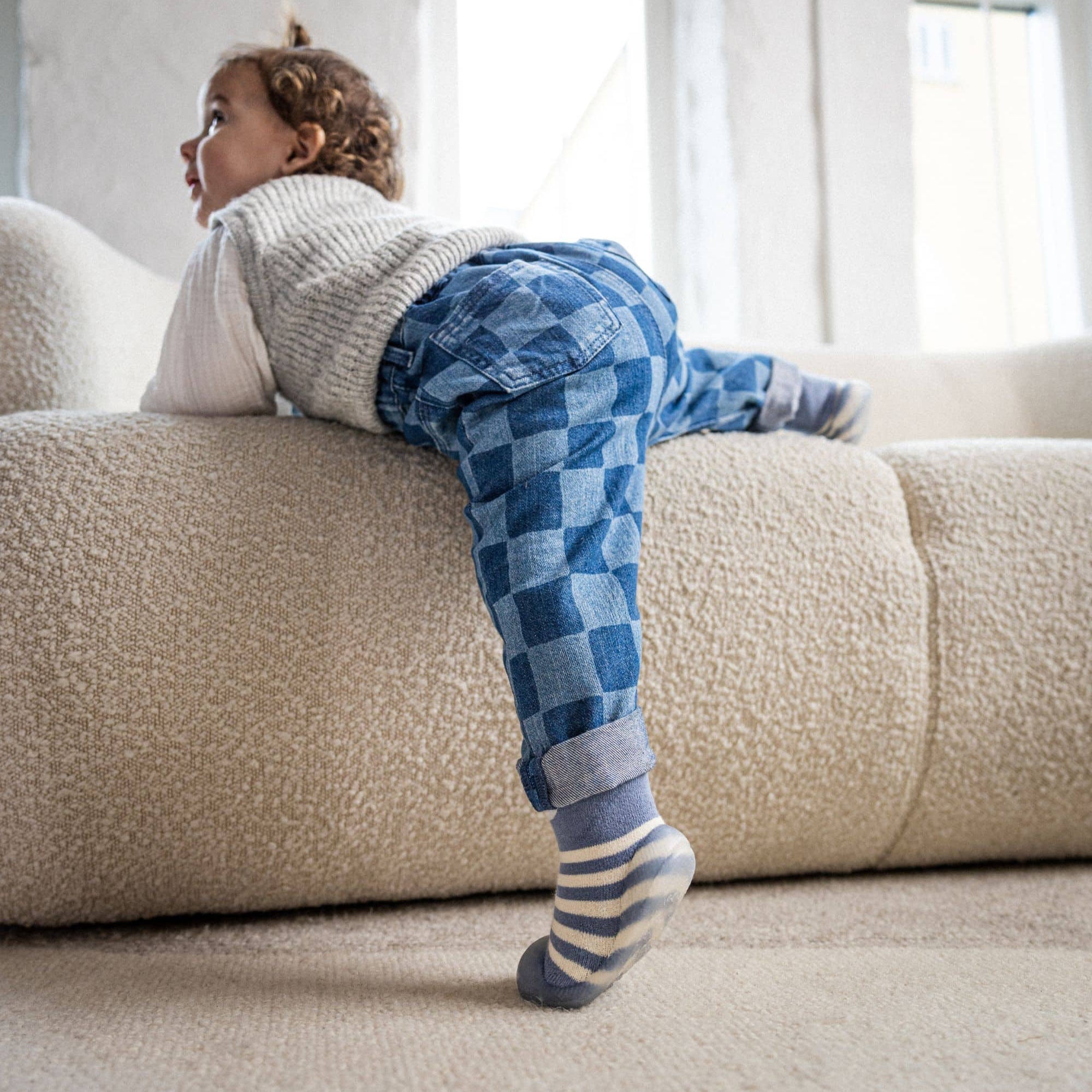 Barn i blå-stribede begyndersko fra Minifus klatrer op ad sofa, iført ternede bukser og strikvest i en lys stue.