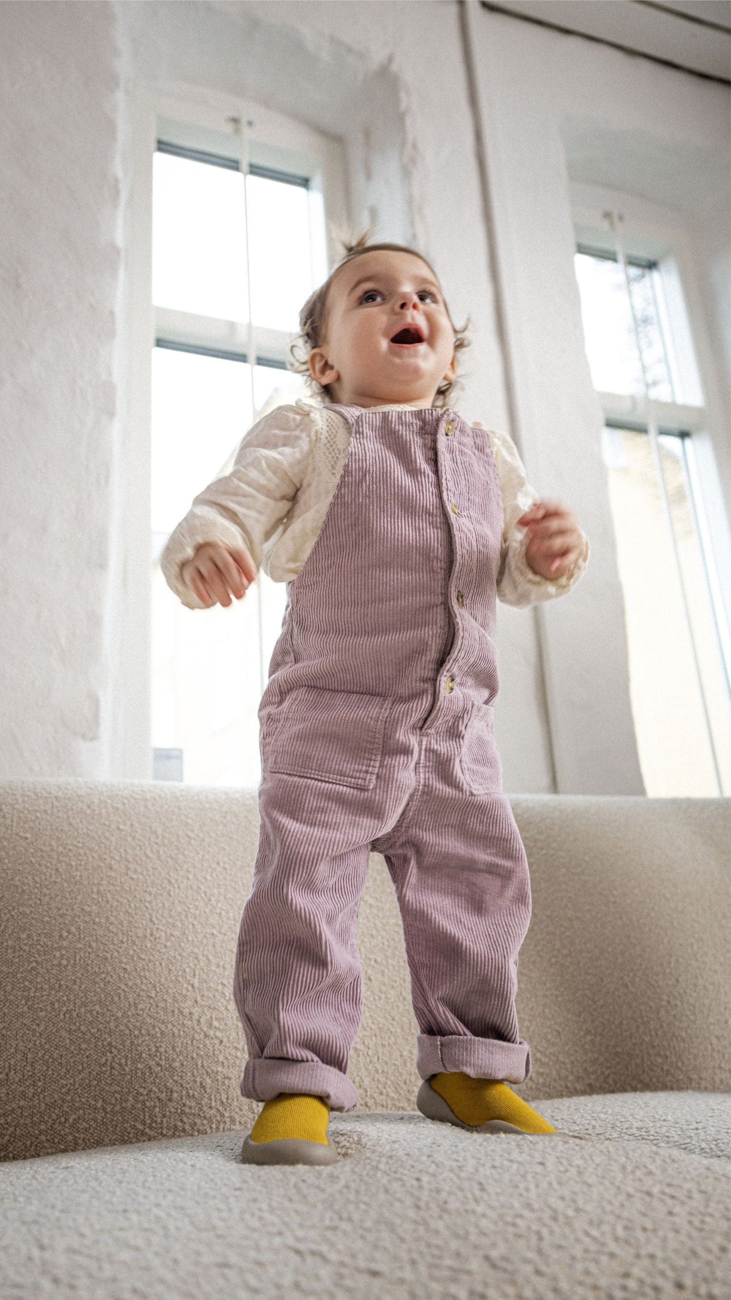 Barn iført gule begyndersko står på sofa med skridsikre sko til leg og balance for de mindste