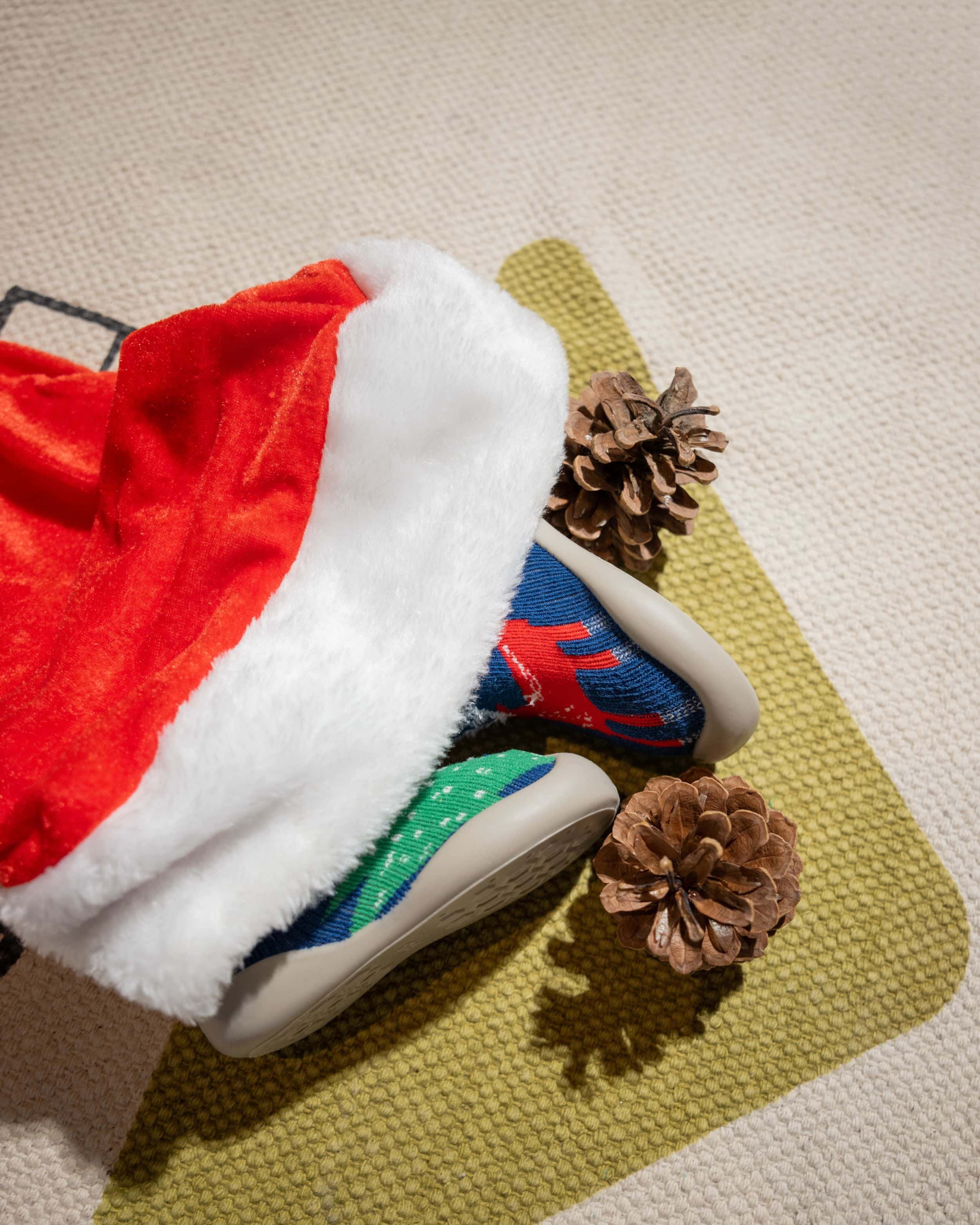 Blå julesko til børn med rensdyrmotiv delvist dækket af nissehue, omgivet af grankogler.