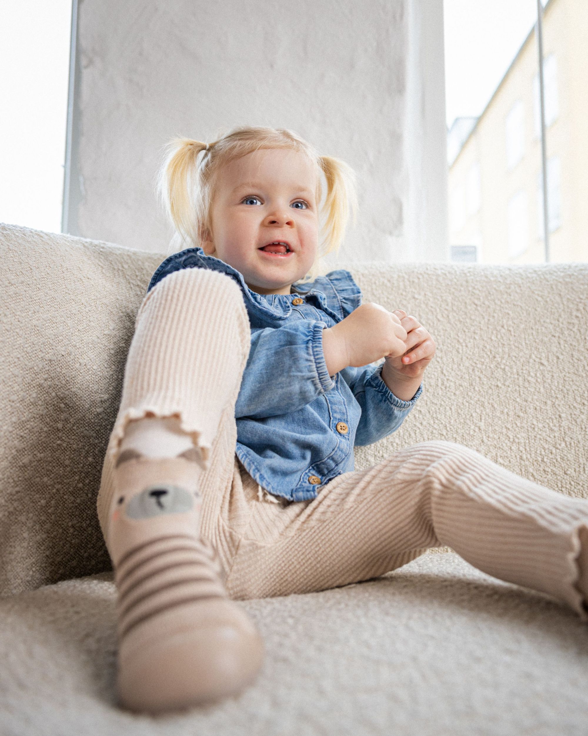 Lille pige iført brun bjørne-begyndersko fra Minifus, siddende afslappet i sofa med et smil og bløde, ribbede leggings.
