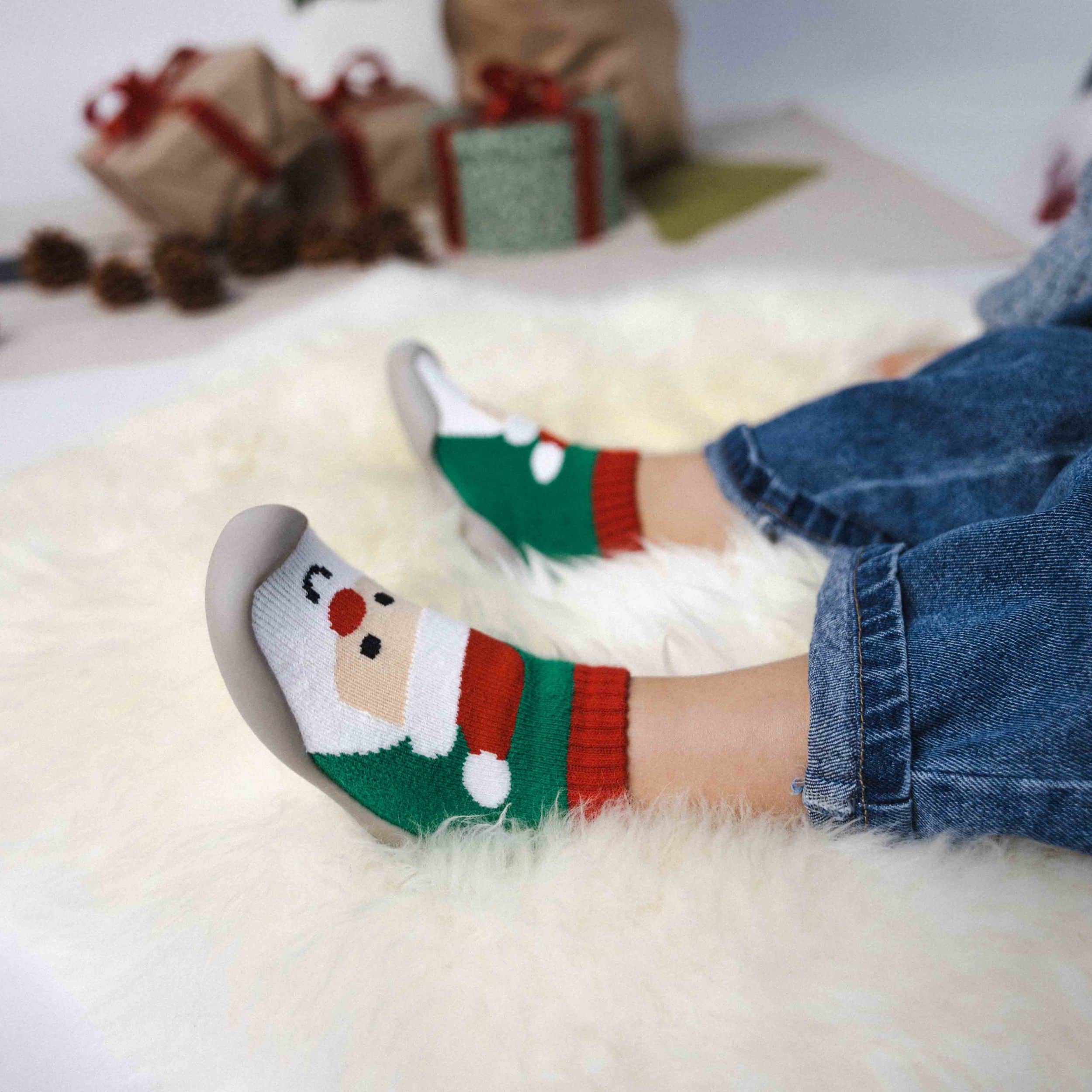 Barn iført børne julesko med julemandmotiv på skridsikker sål på blødt tæppe med julegaver i baggrunden